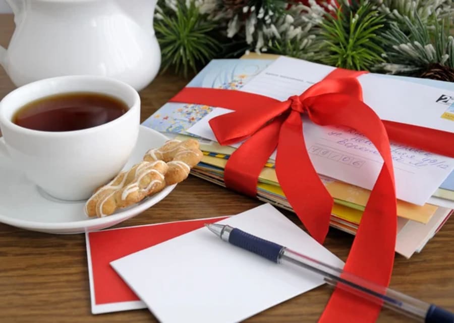 Stack of Christmas cards tied with a red ribbon beside a cup of tea, cookies, a pen and a blank note card.