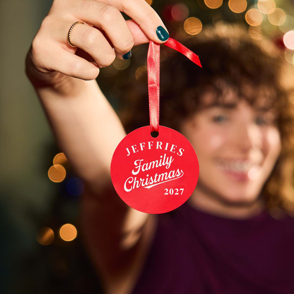 Red personalized Christmas ornament reading “Jeffries Family Christmas 2027” held in front of festive lights.