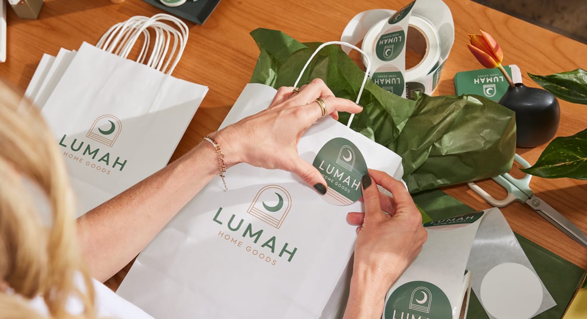 A green and pink logo for a home goods brand featuring a crescent moon on a paper bag and a sticker