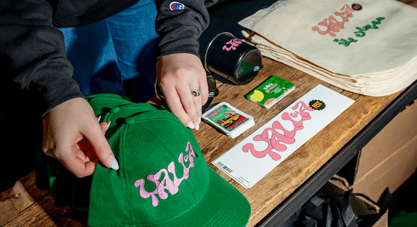 Hands placing a green baseball cap with pink embroidered logo onto a wooden table next to branded merch including stickers, business cards, a mug and tote bags