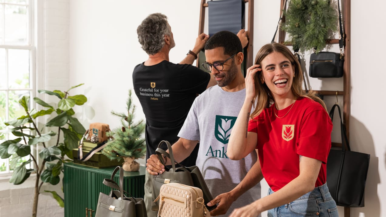 Three employees in a luggage shop wearing different styles of same brand T-shirt: red with logo on front left, light gray with logo and typography, as well as black with logo and “grateful for your business this year” on back center.