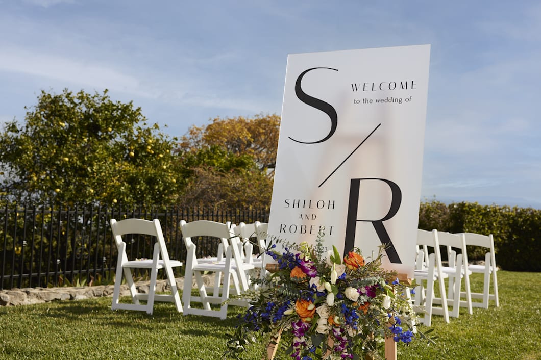 An outdoor wedding with a welcome board, white chairs and flowers
