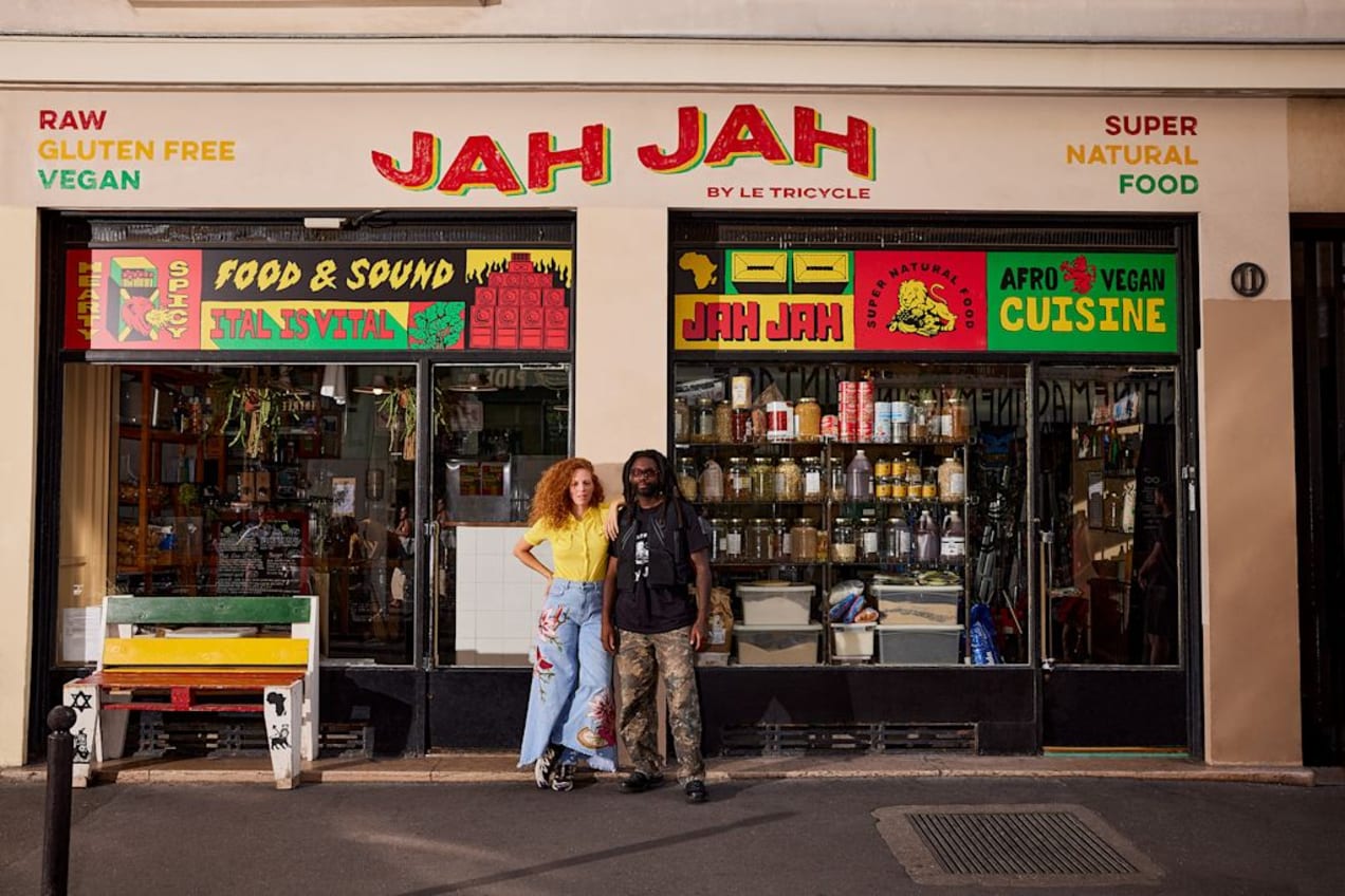 Two small business owners standing outside their natural food store with two large, multicolored banners displayed in the windows