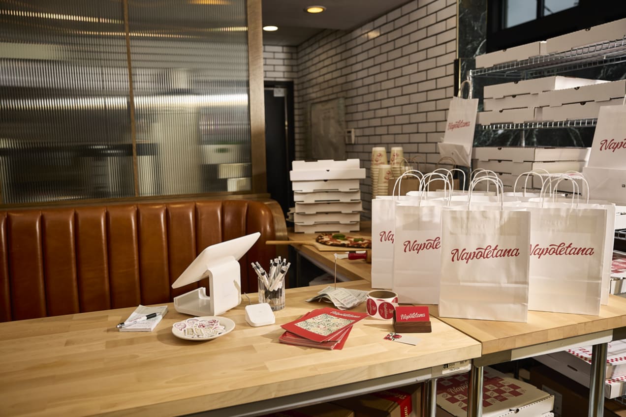 The counter at a pizza restaurant with branded white paper bags, pizza boxes, pens and roll of logo stickers