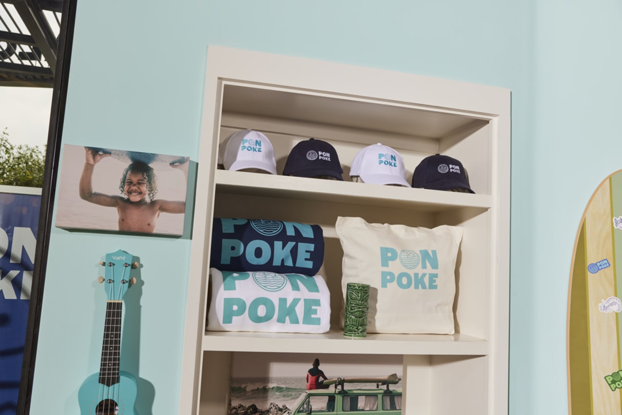 A shelf featuring merch including a tote bag, caps, and folded branded hoodies in white and blue
