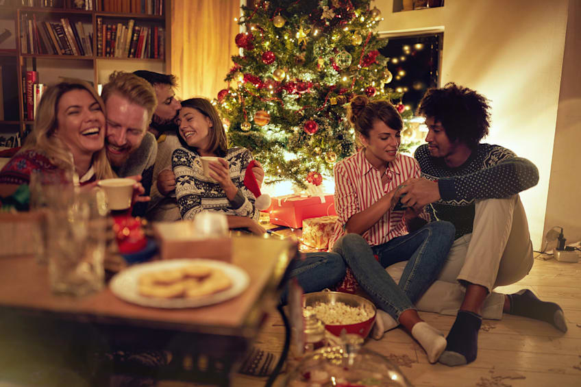Friends enjoying a holiday gathering with drinks and snacks beside a decorated Christmas tree.