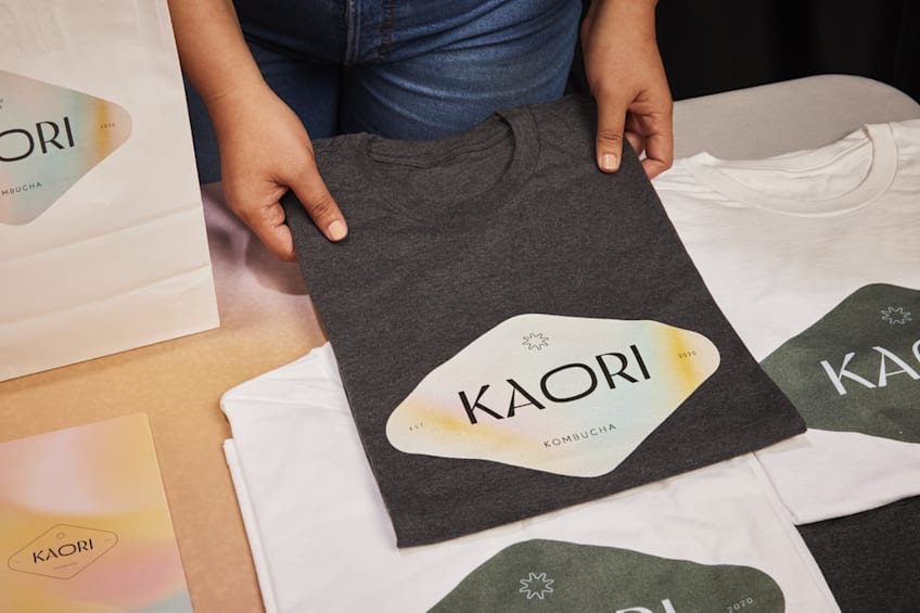 Small business owner arranging white and gray folded T-shirts on a table with diamond shaped “Kaori” logos