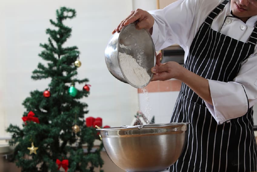 A person pouring caster sugar in a metallic bowl with a Christmas tree standing in the background