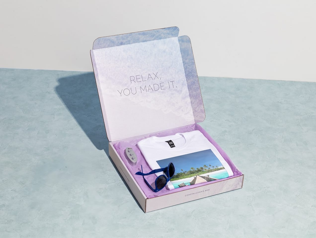 A purple cardboard mailer box containing branded products including a t-shirt and sunglasses.