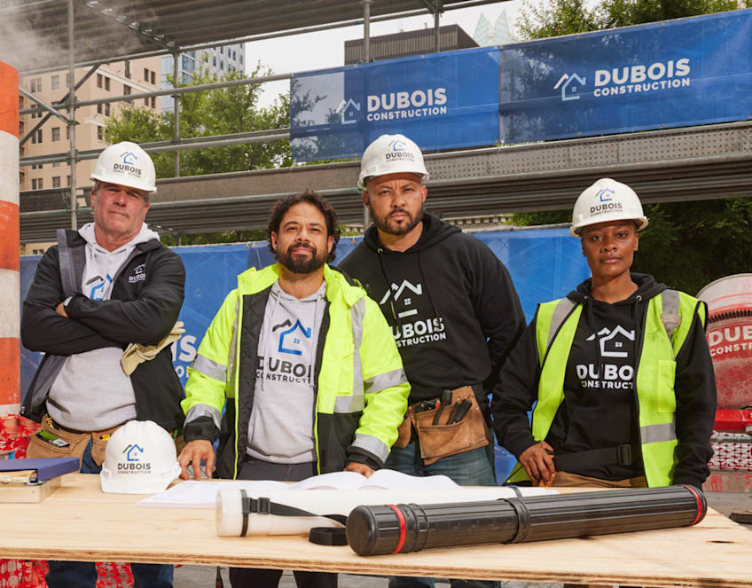 Group of four construction workers on a building site wearing black and gray hoodies with “Dubois Construction” logo.