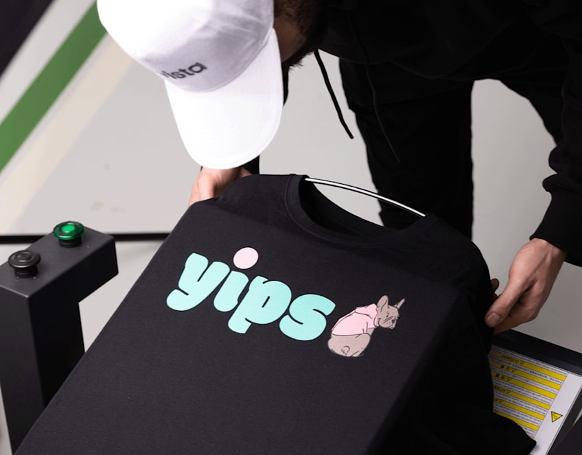Someone taking a black T-shirt out of a DTG printer, printed with a pink dog graphic and baby blue text &ldquo;Yips&rdquo;