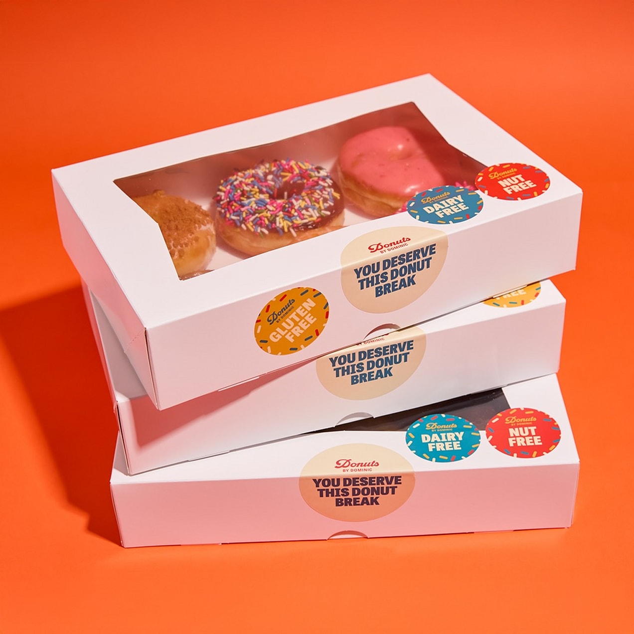 Stacked white bakery boxes with a clear window showing assorted donuts and colorful branded allergen-alert stickers on the lids – “dairy-free,” “nut free,” and “gluten-free”.