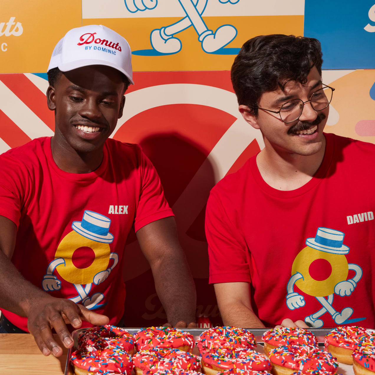 Two donut shop employees behind a counter full of donuts wearing matching red T-shirts with donut graphic and their names &ldquo;Alex&rdquo; and &ldquo;David&rdquo; respectively.
