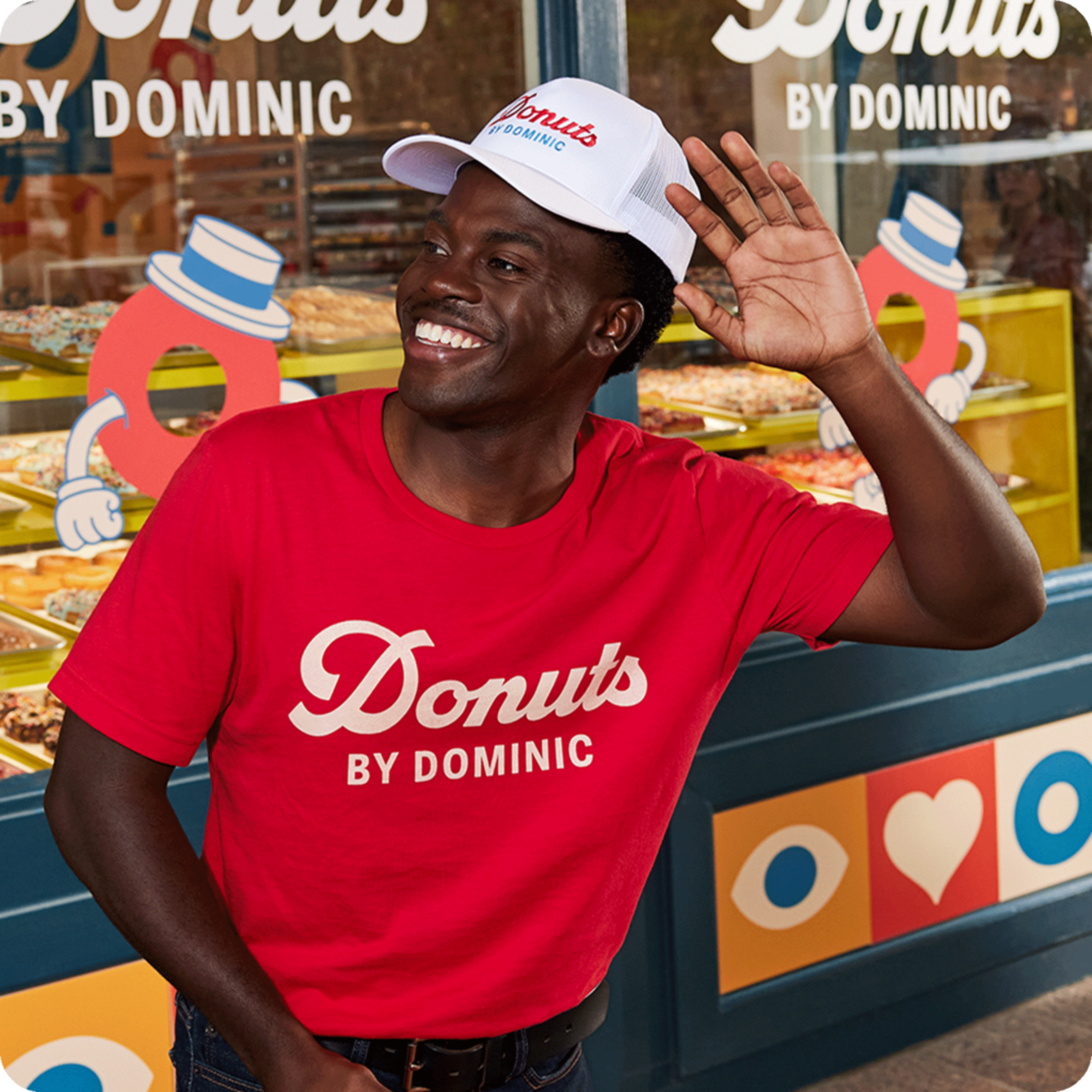 Donut shop employee outside shop wearing white branded trucker cap and red T-shirt with “Donuts” and “By Dominic” in two different brand fonts
