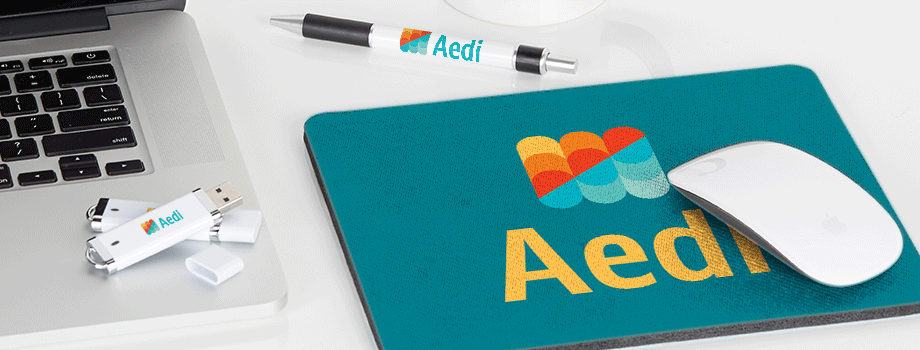 Branded merchandise on a white desk including a mug, USB stick, pen, and mousepad printed with a colorful logo