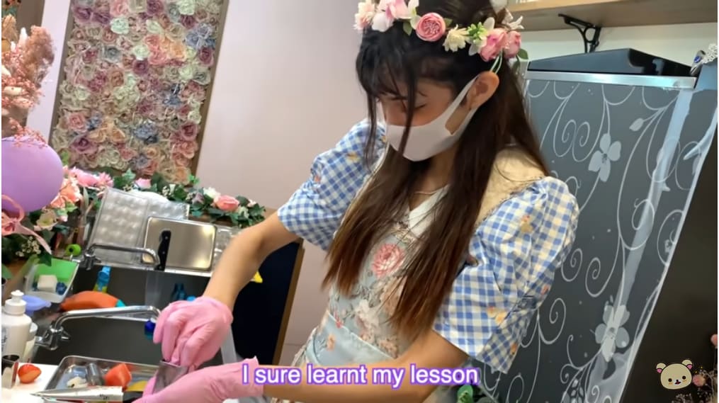  A lady dressed in blue plaid dress and floral apron is seen preparing skewered fruits
