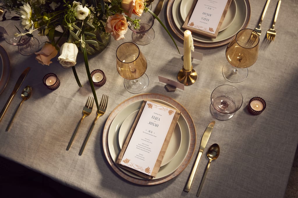 a wedding table setup with matching cutlery, crockery, candles, flowers, and a personalized menu card
