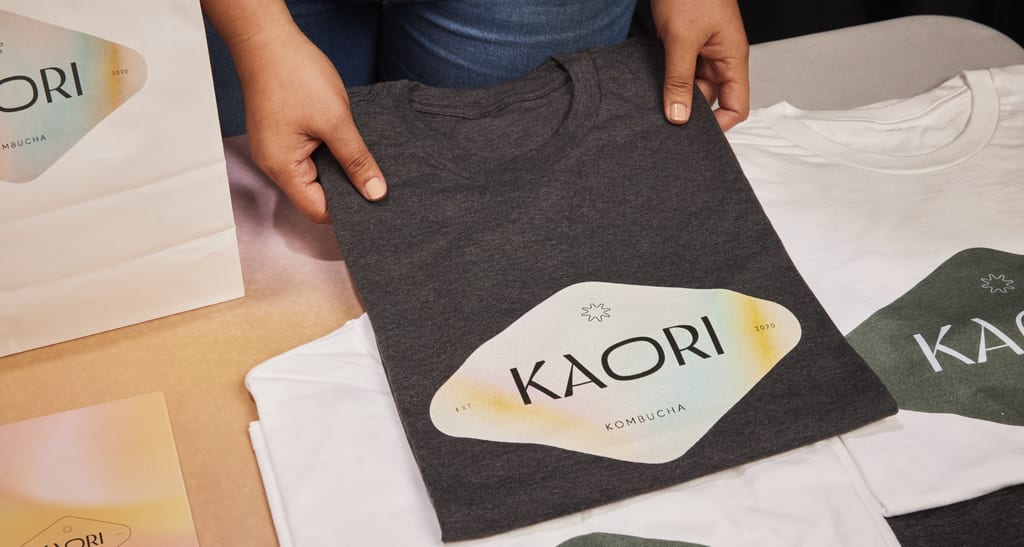 Folded t-shirts in different colors with logo variations, displayed on a table, with hands presenting a black t-shirt with a light-colored logo.