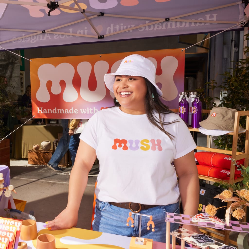 Small business owner stood behind a stall at a trade show wearing white T-shirt with orange, purple, yellow, pink colorful font “MUSH.”