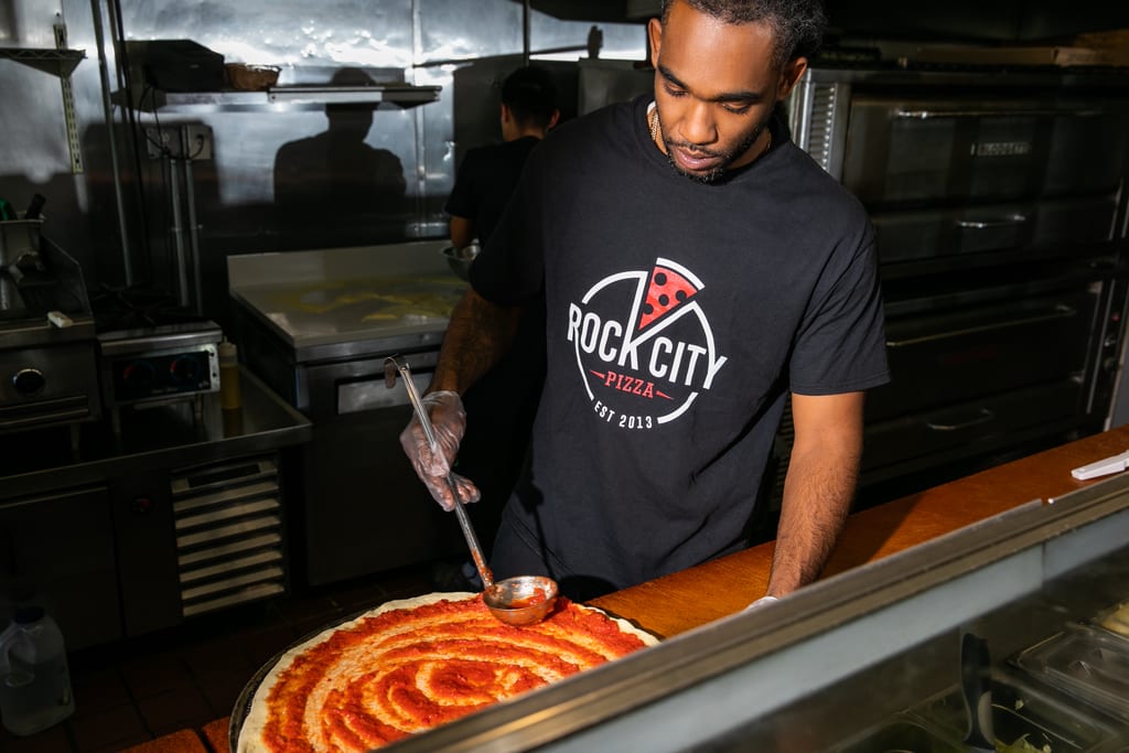 Man in a kitchen spreading tomato sauce on a pizza base wearing black-T-shirt with red and white circular pizza graphic for “ROCK CITY PIZZA.”