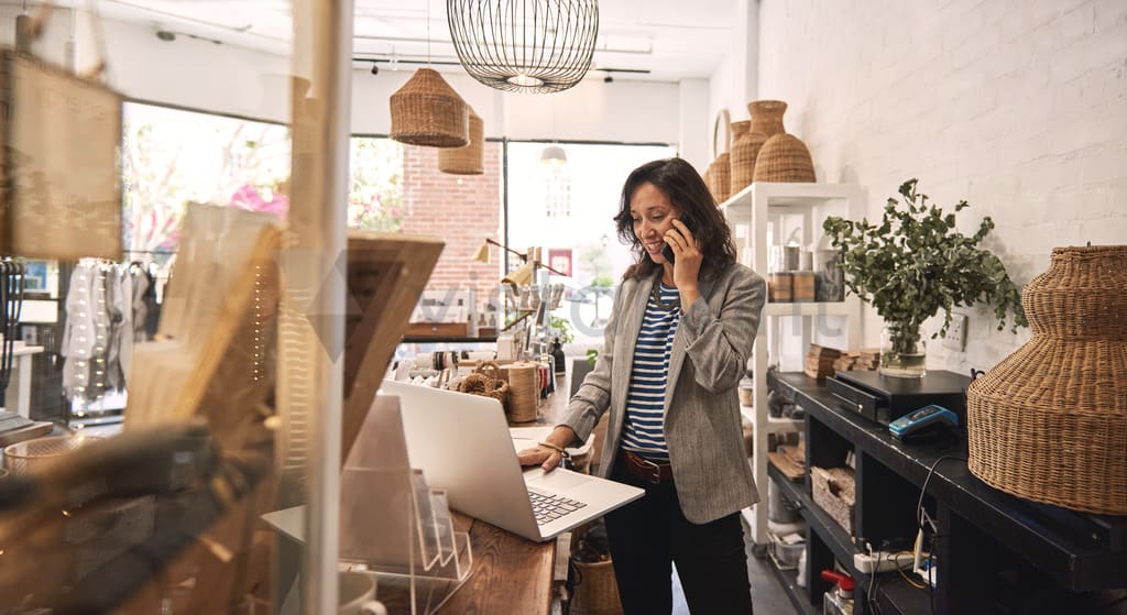 Person on phone behind the counter of a homestore and also operating a laptop.