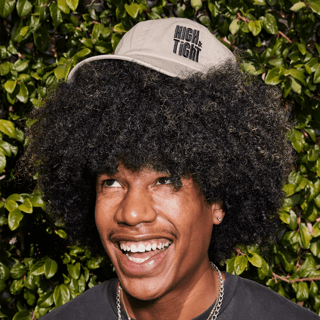 Joyful man with tall curly hair, playfully poses with an embroidered brand hat balanced on his hair.