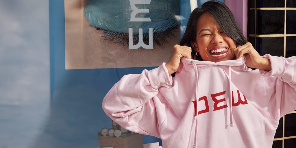 Small business owner in front of store pulling up a baby pink hoodie with red text &ldquo;DEW&rdquo; across the chest.