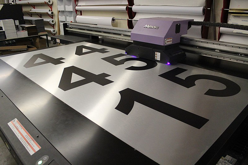 Large-format printer printing black numbers on a brushed aluminum sheet in process.