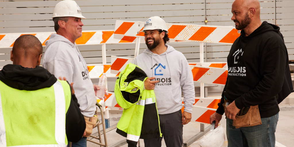 Four construction workers on a building site wearing branded hoodies, two in black two in light grey, with matching house graphic logo and &ldquo;DUBOIS CONSTRUCTION&rdquo; text.