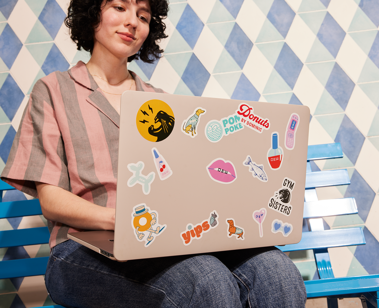 A woman sitting in front of a wall with a white and blue diamond design and using a laptop with stickers on the lid