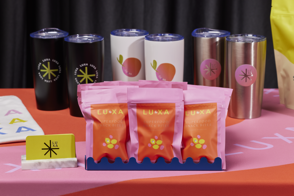 LUXA branded superfood snack bags and reusable tumblers displayed on a colorful promotional table.