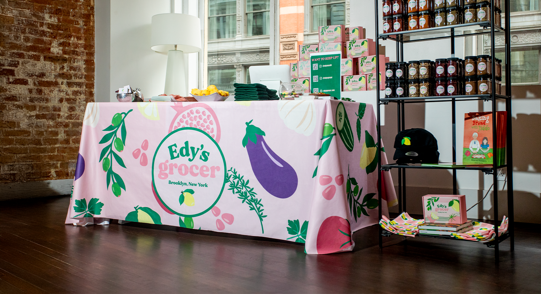 Edy’s Grocer display with a pink branded tablecloth, packaged products, hats, books, QR code poster, and jars on shelving.
