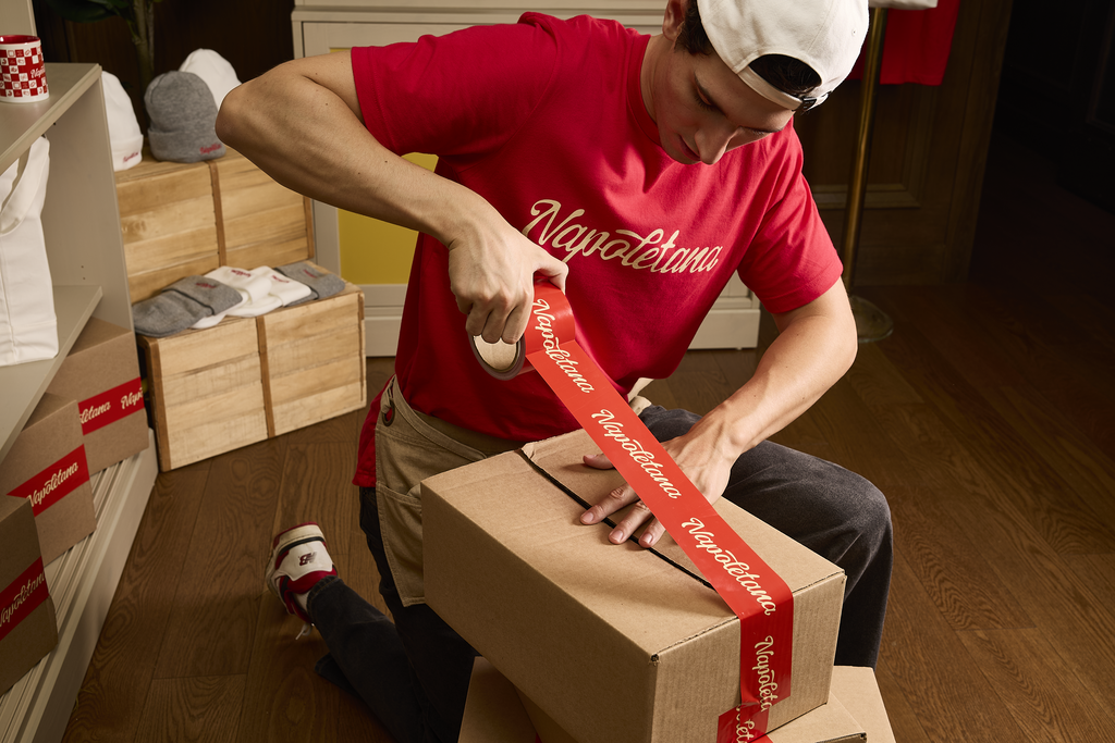 Small business owner taping large cardboard boxes with red custom packing tape with white repeating “Napoletana” text