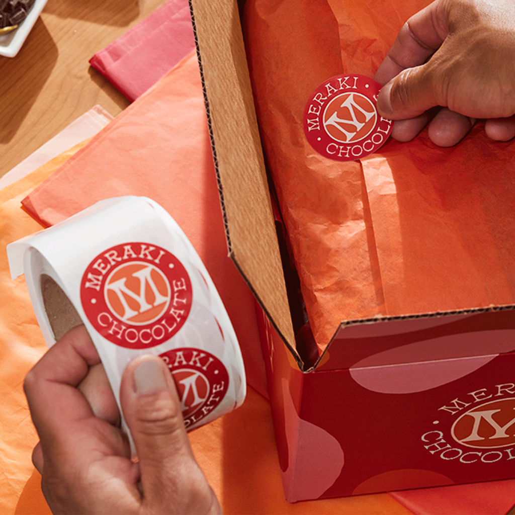 Small business owner placing branded circular logo sticker onto orange inner tissue inside a custom red package