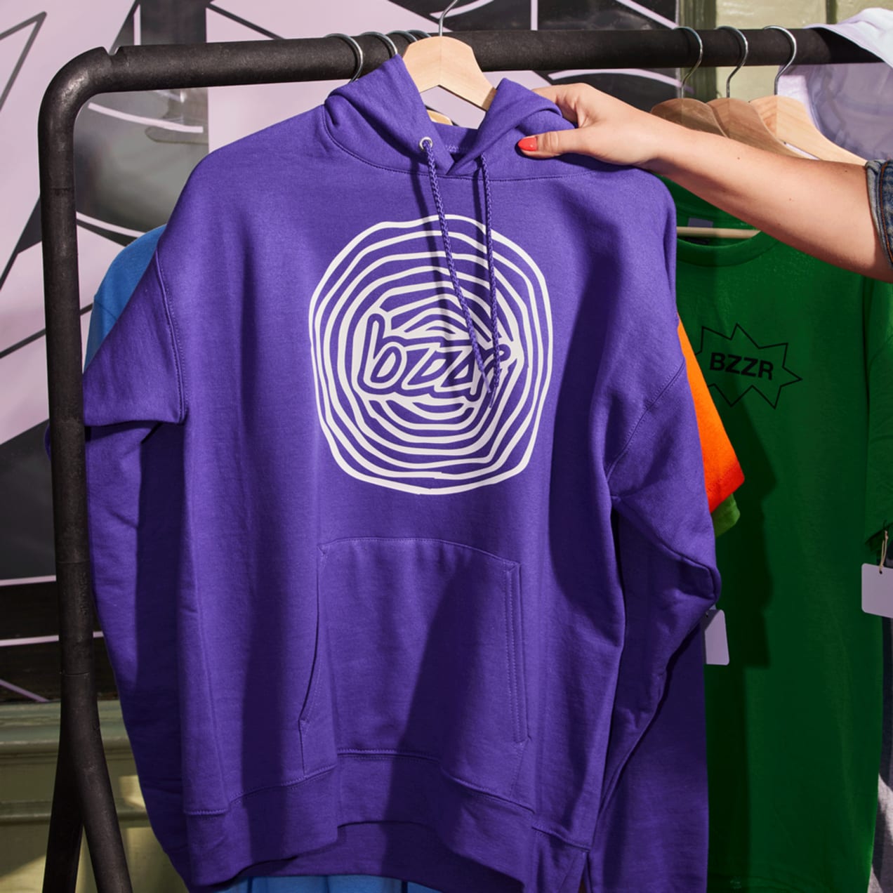 Hand reaching out for a purple hoodie on a rack with a white spiral design logo with “bzzr” at the center.