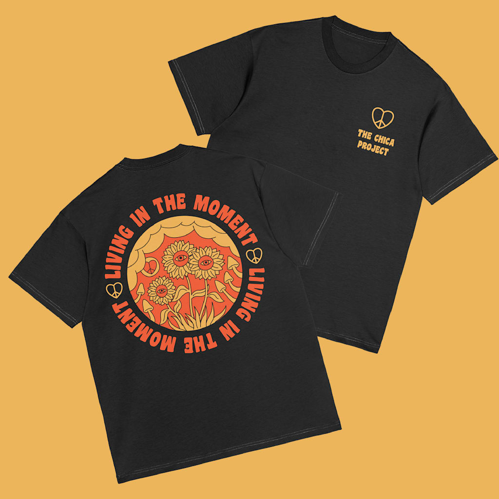 Front and back of black T-shirt with small orange peace heart logo and font “The Chica Project on front left and large orange circular floral graphic with “LIVING IN THE MOMENT” around it.