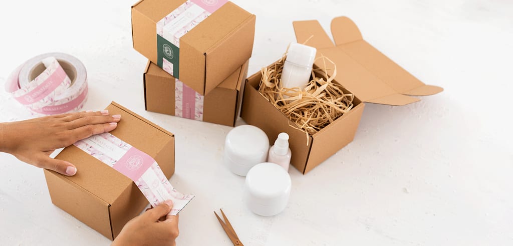 Hands packaging self-care products in carton boxes with branded sticker seals in delicate pinks and white