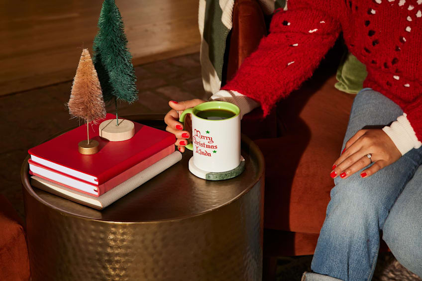 Woman in red sweater leaning over a festive side table with holiday decor to grab a personalized holiday mug that says “Merry Christmas, Linda.”