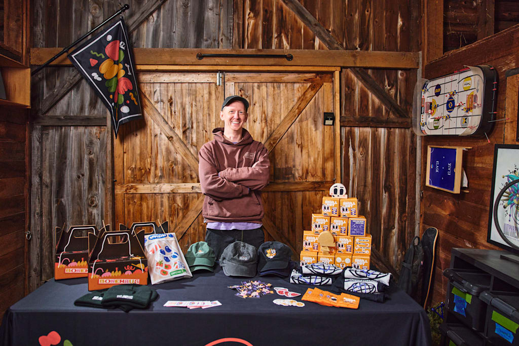 Direct photograph of the featured artist/designer, Kirk Wallace, standing behind a table featuring multiple example of his work with VistaPrint products eg. apparel, mugs, packaging, stickers
