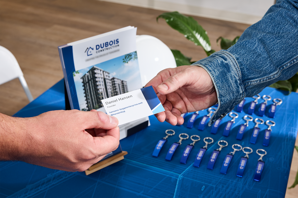 Small business owner handing a business card to another person at a trade show table with Dubois Construction leaflets and blue promotional keyrings.