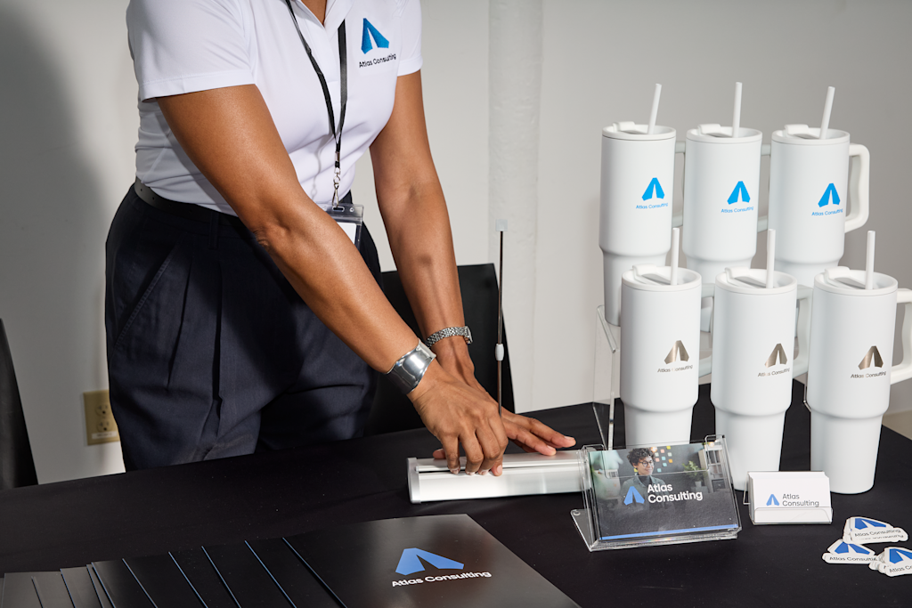 A trade show display table featuring Atlas Consulting-branded tumblers, a business card holder and promotional materials.