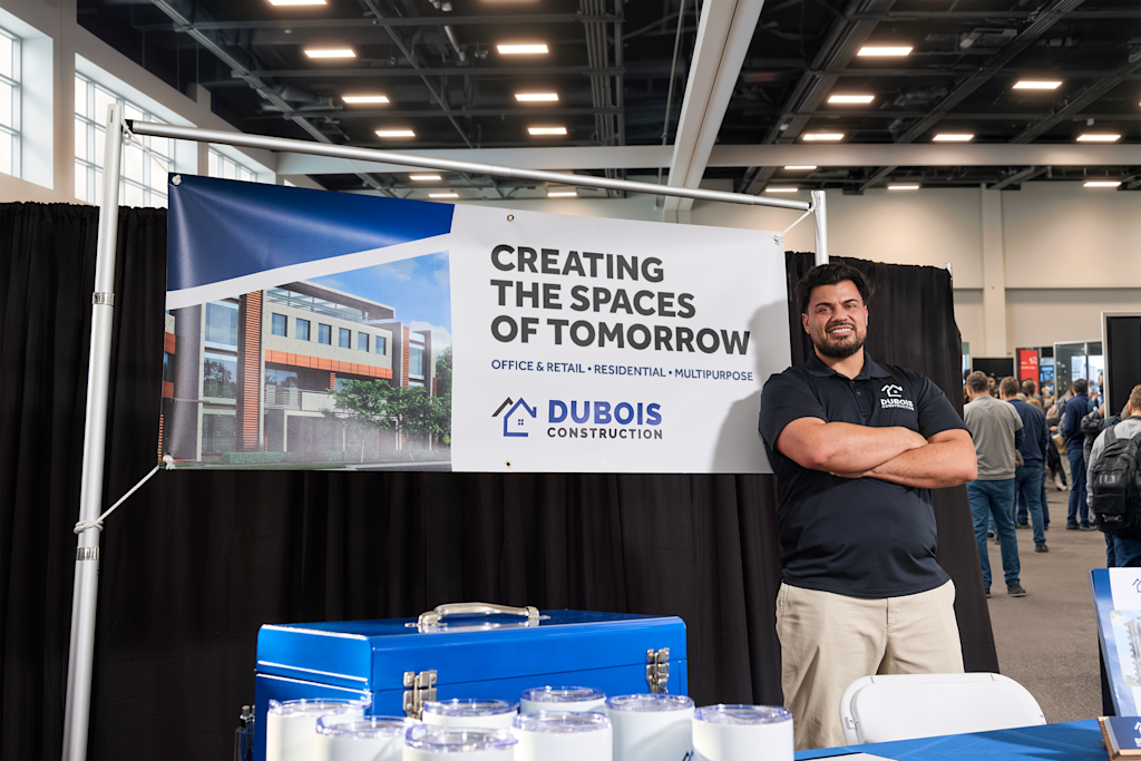 A person standing at a construction company trade show booth, with a branded banner behind them promoting office, retail, and residential construction services.