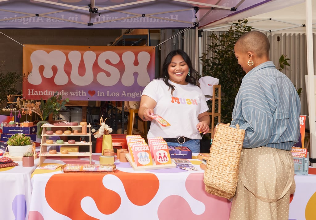 Jewellery vendor serves customer at a well-branded market stall featuring organic shapes and a custom logo across a sunset-coloured banner and tablecloth.