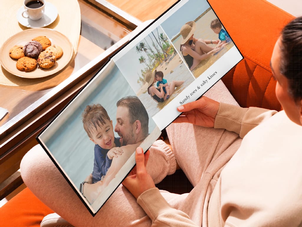 Woman sat on orange couch holding open a custom photo book with images of family on holiday at the beach.