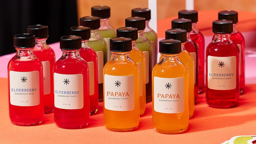 Minimalist beverage packaging featuring small glass bottles with black caps and clean white labels, showcasing brightly colored juices in red, orange and green tones.