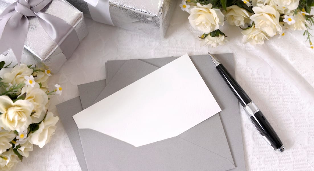 Writing paper or wedding invitation with envelope laid on bridal lace with several wedding gifts and white rose bouquets. Space for copy.