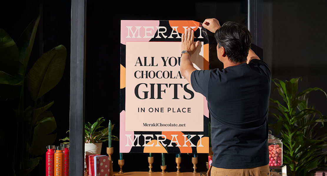 Store owner applying adhesive poster to storefront window surrounded by branded products and potted plants — signage for business in action.