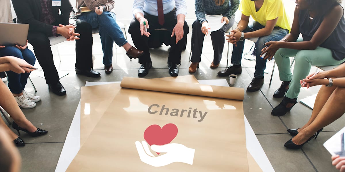 Multicultural group sitting in a circle around a large charity poster with a hand and heart logo.
