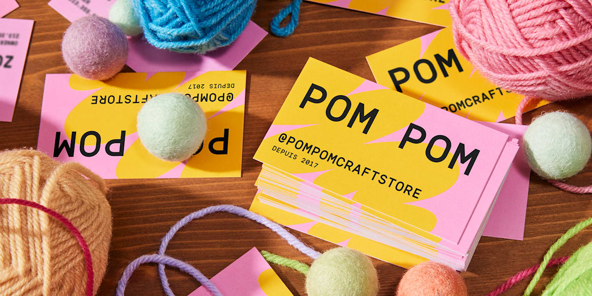 Cartes de visite colorées de la marque PomPom, éparpillées sur une table avec des pelotes de laine – une source d’inspiration pour concevoir une carte de visite.