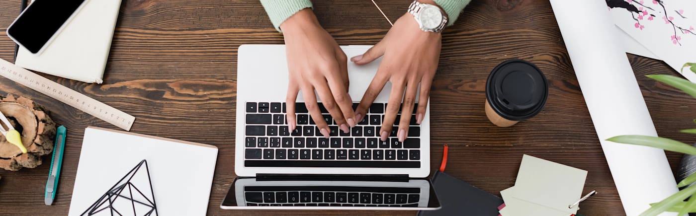 Hands typing on a laptop surrounded by stationery, coffee cup, and smartphone – web pages design software in use.